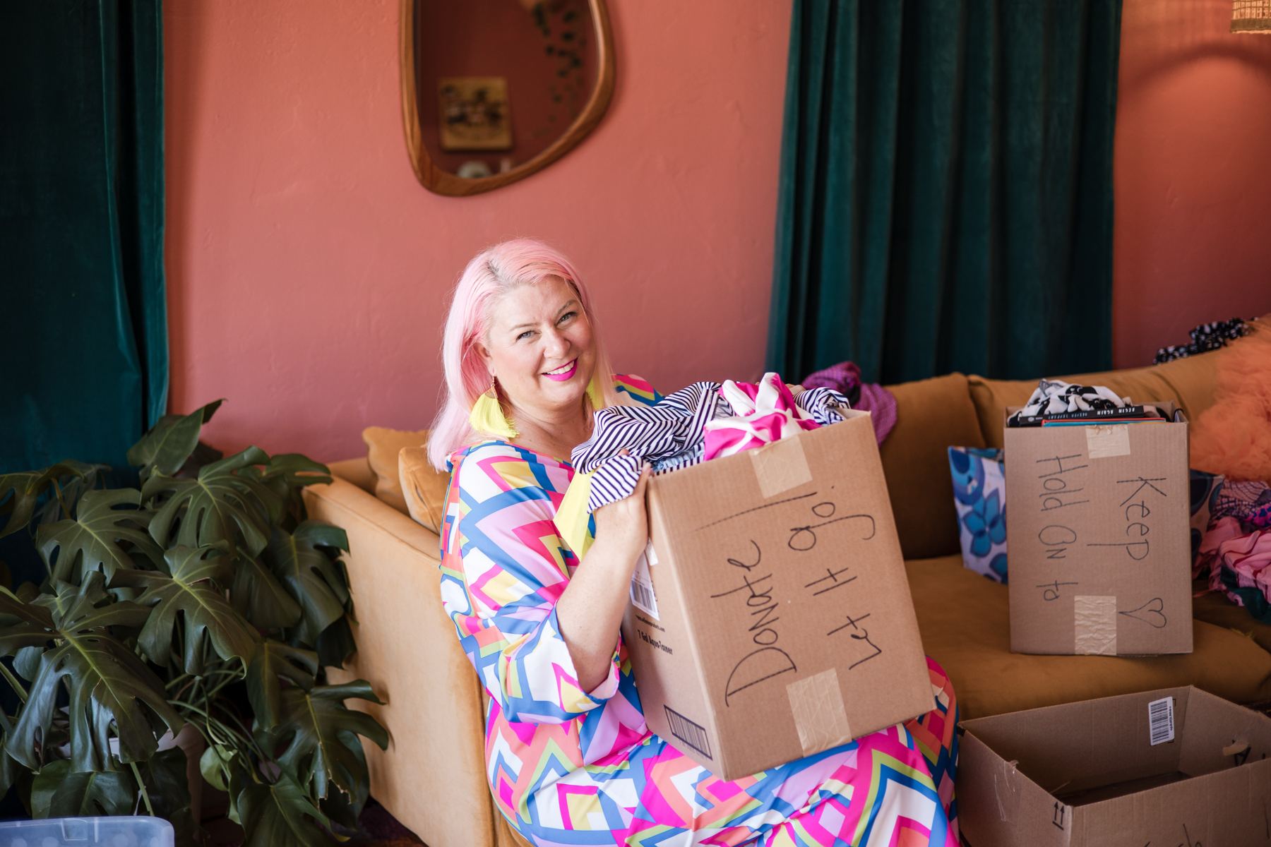 Host Unison Frances, with pink hair, smiling on a couch while holding a box labeled Donate, Let it go.